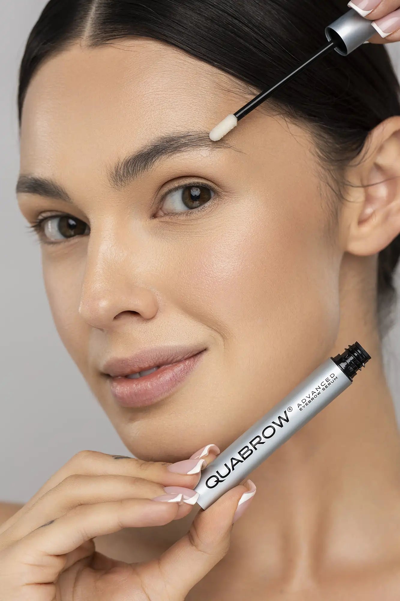Brunette 여성이 애플리케이터 팁이 클로즈업된 QUABROW Advanced Eyebrow Serum을 눈썹 부위에 바르는 모습